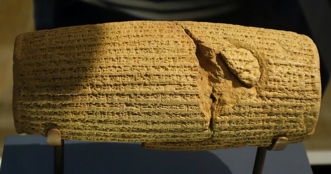A clay cylinder covered in rows of cuneiform, on a museum mount.